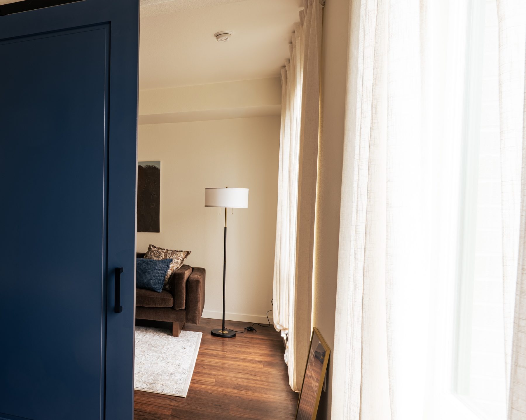 View through the bedroom doorway into the living room with a floor lamp and sheer curtains